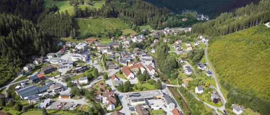 Luftaufnahme eines kleinen Dorfes zwischen grünen Hügeln, mit einer Mischung aus Wohnhäusern, Bäumen und einer gewundenen Straße. Der Himmel ist teilweise bewölkt.