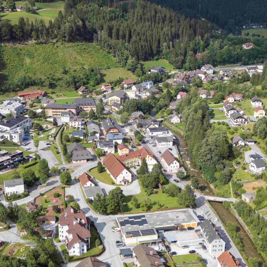 Luftaufnahme eines kleinen Dorfes zwischen grünen Hügeln, mit einer Mischung aus Wohnhäusern, Bäumen und einer gewundenen Straße. Der Himmel ist teilweise bewölkt.