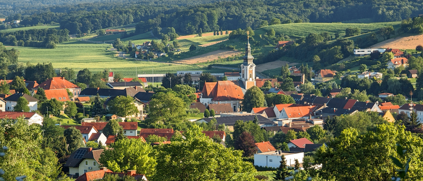 Eine Stadt mit zahlreichen Gebäuden, darunter Häuser und eine Kirche, liegt eingebettet zwischen üppigen grünen Hügeln und Bäumen. Die Landschaft ist mit Feldern durchsetzt, und Berge sind in der Ferne sichtbar.