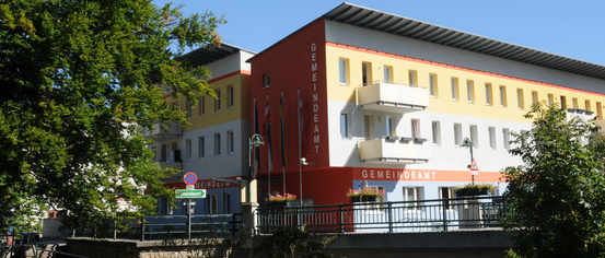 Ein mehrstöckiges Gebäude mit Fahnen davor. Das Wort 'GEMEINDEAM' steht in Rot auf dem Gebäude. Auf beiden Seiten des Gebäudes stehen zwei Lampen. Ein grünes Straßenschild steht vor dem Gebäude.