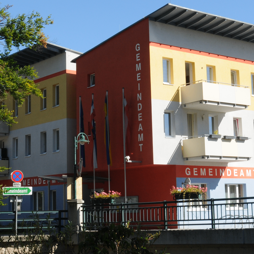 Ein mehrstöckiges Gebäude mit Fahnen davor. Das Wort 'GEMEINDEAM' steht in Rot auf dem Gebäude. Auf beiden Seiten des Gebäudes stehen zwei Lampen. Ein grünes Straßenschild steht vor dem Gebäude.