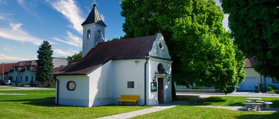 Eine kleine weiße Kirche mit einem braunen Dach und einem Glockenturm steht unter einem blauen Himmel mit Wolken. Ein gelber Stuhl ist vor der Kirche platziert.