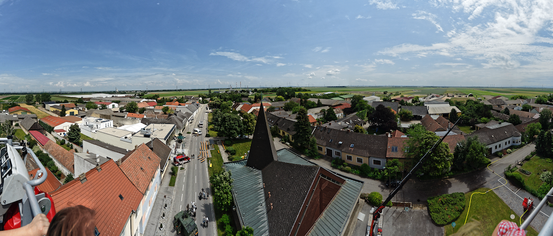 Luftaufnahme einer Stadt mit einer Hauptstraße, Gebäuden und einer Kirche mit einem Turm. Der Himmel ist blau mit verstreuten Wolken.