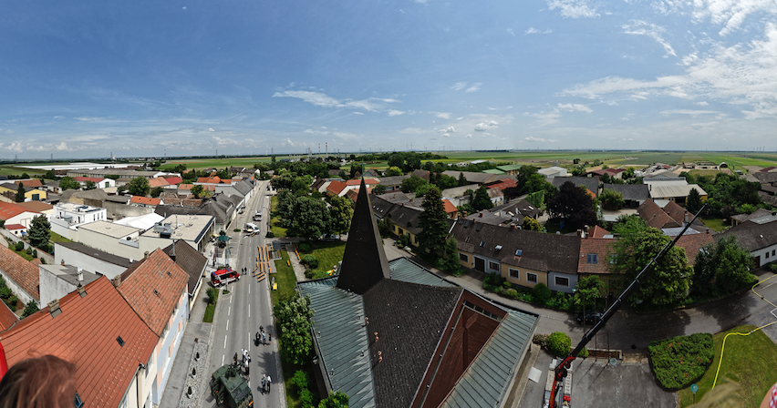 Luftaufnahme einer Stadt mit einer Hauptstraße, Gebäuden und einer Kirche mit einem Turm. Der Himmel ist blau mit verstreuten Wolken.