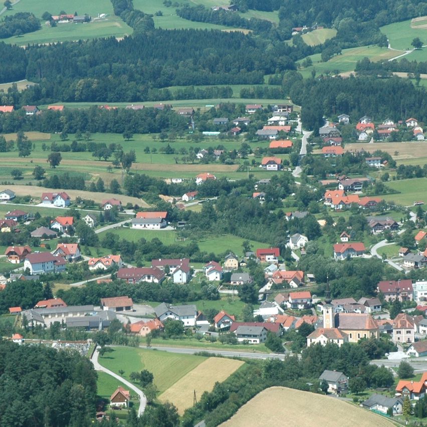 Ein Luftbild eines kleinen Dorfes, umgeben von üppigen grünen Feldern und dichten Wäldern. Das Dorf besteht aus zahlreichen Häusern mit roten Dächern, eingebettet zwischen Bäumen.