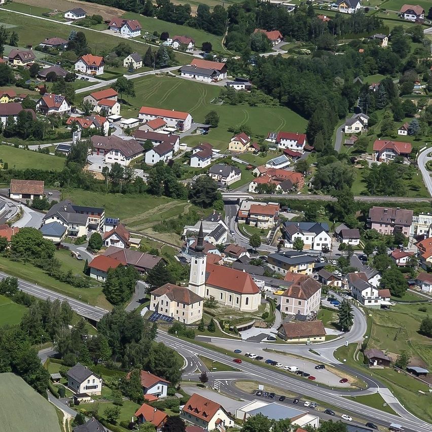 Luftaufnahme eines ländlichen Dorfes mit vielen Häusern, einige mit roten Dächern, eingebettet in eine üppige grüne Landschaft. Das Dorf ist von landwirtschaftlichen Flächen und Bäumen umgeben, mit einer zentralen Kirche und einer kurvenreichen Straße, die hindurchführt.