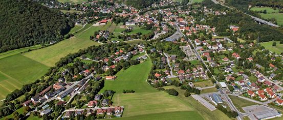 Luftaufnahme einer ländlichen Stadt, umgeben von üppigen grünen Feldern und Hügeln unter einem klaren blauen Himmel. Viele Häuser, meist mit roten Dächern, sind entlang der gewundenen Straßen zu sehen.