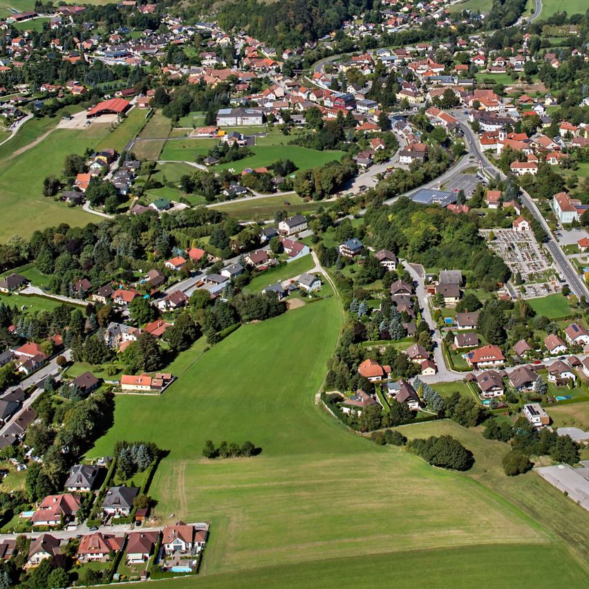 Luftaufnahme einer ländlichen Stadt, umgeben von üppigen grünen Feldern und Hügeln unter einem klaren blauen Himmel. Viele Häuser, meist mit roten Dächern, sind entlang der gewundenen Straßen zu sehen.