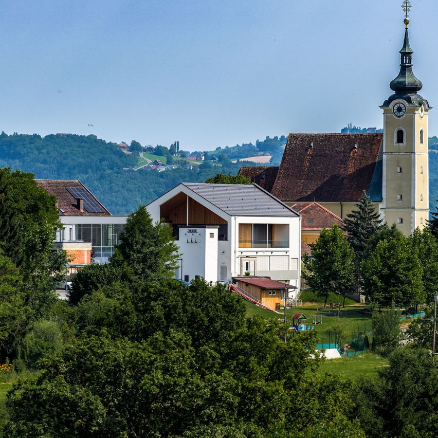 Eine Stadtlandschaft zeigt einen hohen Glockenturm und eine Kirche, umgeben von üppigem Grün und Bäumen unter einem klaren blauen Himmel.