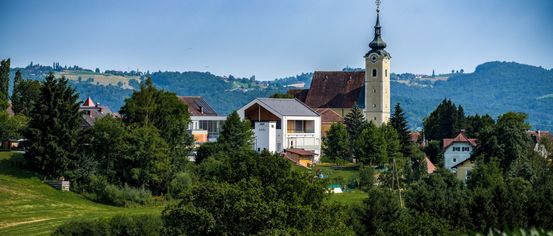 Eine Stadtlandschaft zeigt einen hohen Glockenturm und eine Kirche, umgeben von üppigem Grün und Bäumen unter einem klaren blauen Himmel.