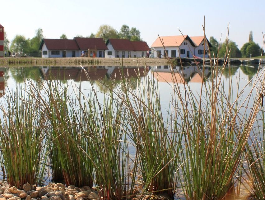 Ein ruhiger See mit hohen Gräsern im Vordergrund. Im Hintergrund befinden sich einige Häuser, eines mit einem roten Dach, und ein kleiner rot-weißer Leuchtturm. Bäume und eine Brücke sind in der Ferne sichtbar.