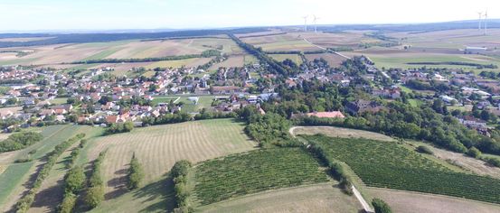 Luftaufnahme eines Dorfes mit mehreren Häusern, einige mit roten Dächern, umgeben von grünen Feldern und Bäumen. In der Ferne stehen zwei Windturbinen in der Nähe einer Straße.