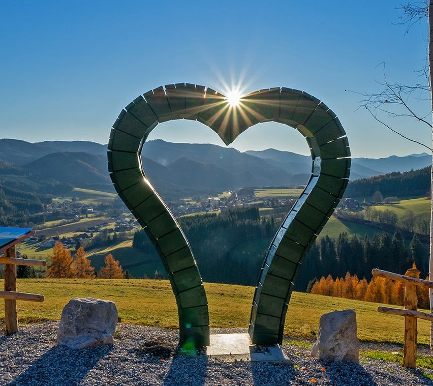 Eine grüne Herzskulptur auf einem Hügel mit klarem blauem Himmel und Bergen im Hintergrund. Ein Dorf ist im Tal unten sichtbar.