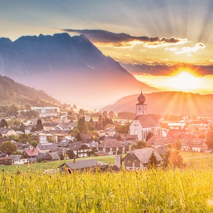 Sonnenuntergang über einer kleinen Stadt mit einer Kirche im Vordergrund, Bergen im Hintergrund und einem klaren Himmel.
