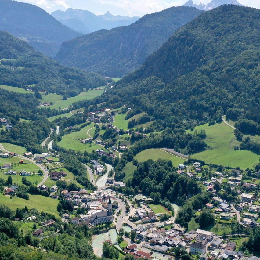 Luftaufnahme eines Tals mit Bergen, grünen Feldern und einer kleinen Stadt. Die Stadt liegt zwischen Hügeln und ist von üppigem Grün umgeben. In der Ferne fließt ein Fluss durch das Tal.