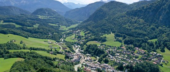 Luftaufnahme eines Tals mit Bergen, grünen Feldern und einer kleinen Stadt. Die Stadt liegt zwischen Hügeln und ist von üppigem Grün umgeben. In der Ferne fließt ein Fluss durch das Tal.