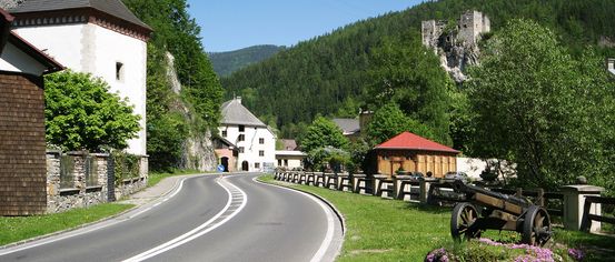 Eine malerische Straße mit weißen Markierungen schlängelt sich durch ein Dorf. Im Hintergrund steht ein Schloss auf einem Hügel. Bäume und Häuser sind entlang der Straße sichtbar.