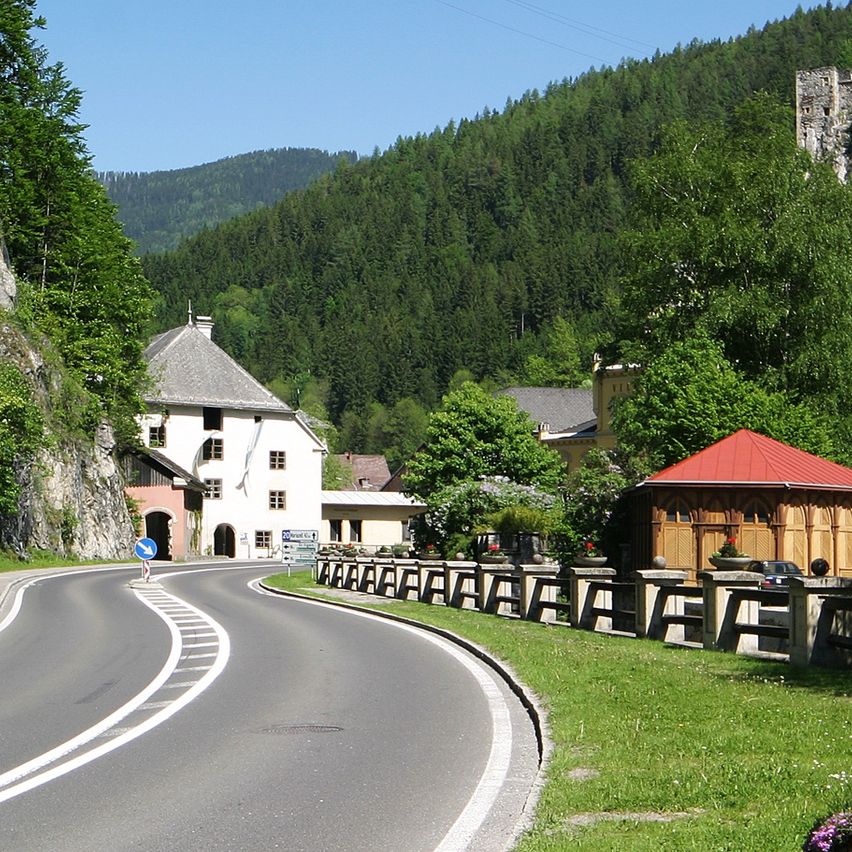 Eine malerische Straße mit weißen Markierungen schlängelt sich durch ein Dorf. Im Hintergrund steht ein Schloss auf einem Hügel. Bäume und Häuser sind entlang der Straße sichtbar.