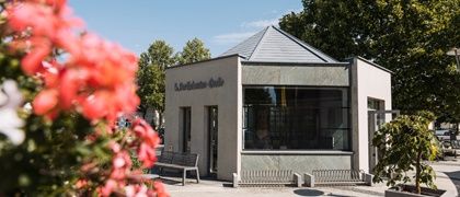 Ein kleines Gebäude mit einem grauen Dach und Glasfenstern ist von Blumen und Bäumen umgeben. Draußen steht eine Bank.