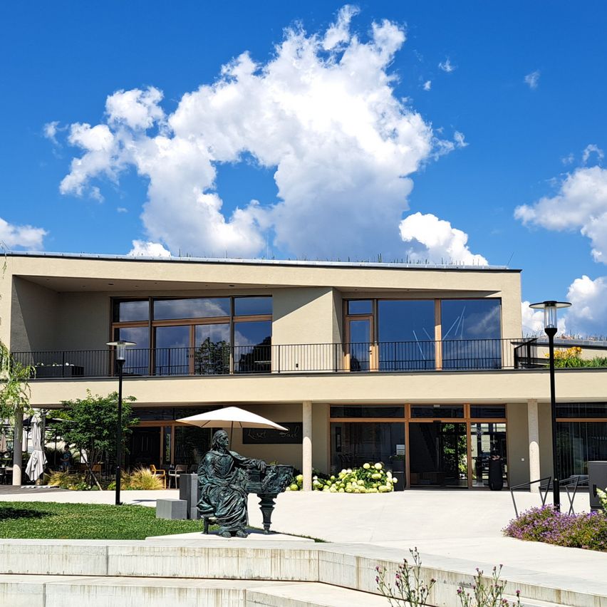 Ein modernes Gebäude mit großen Glasfenstern steht in einem üppigen Garten unter einem teilweise bewölkten Himmel. Eine Statue und ein Regenschirm sind in der Nähe des Eingangs platziert.