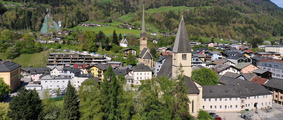 Eine Stadt mit mehreren Häusern und Gebäuden, umgeben von grünen Hügeln und Bergen. Eine Kirche mit zwei Türmen sticht in der Mitte hervor.