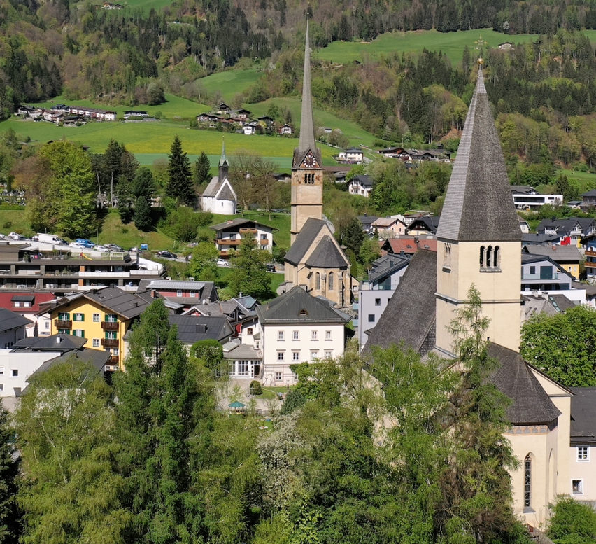 Eine Stadt mit mehreren Häusern und Gebäuden, umgeben von grünen Hügeln und Bergen. Eine Kirche mit zwei Türmen sticht in der Mitte hervor.