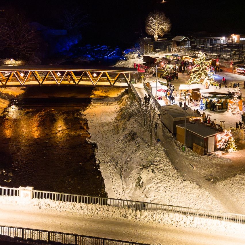 Eine Nachtansicht eines Dorfes mit einer Brücke über einem Fluss, beleuchtet von Lampen. Viele Menschen schlendern auf einem Weihnachtsmarkt am Flussufer entlang.