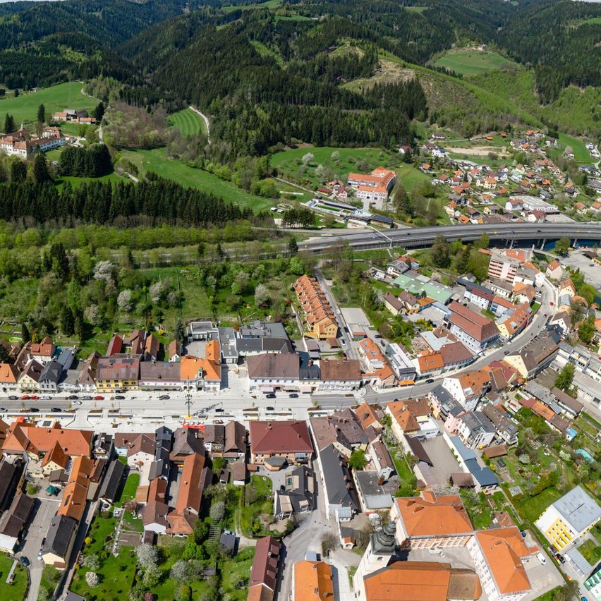 Luftaufnahme einer Stadt mit Gebäuden, Straßen und einer Brücke. Grüne Felder, Bäume und Berge im Hintergrund.