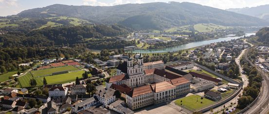 Luftaufnahme einer Stadt mit Gebäuden, einem Fluss und Bergen im Hintergrund. Die Stadt ist von Bäumen und Gras umgeben.