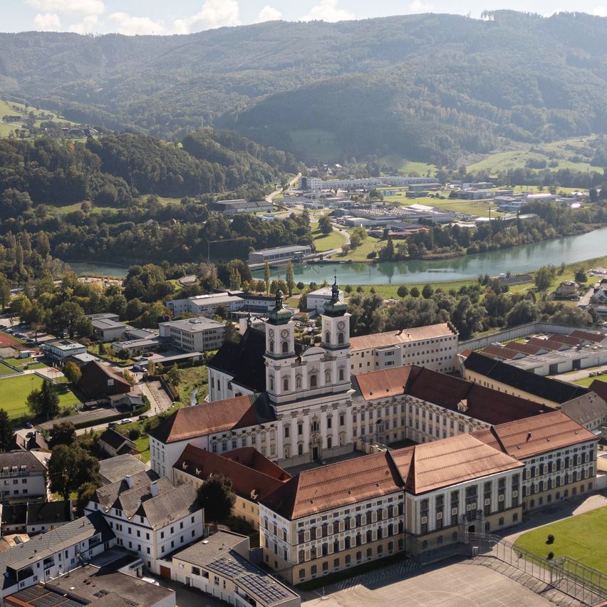 Luftaufnahme einer Stadt mit Gebäuden, einem Fluss und Bergen im Hintergrund. Die Stadt ist von Bäumen und Gras umgeben.