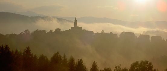 Eine neblige Morgenlandschaft mit einem hohen Turm und Bergen in der Ferne, getaucht im goldenen Licht der aufgehenden Sonne.