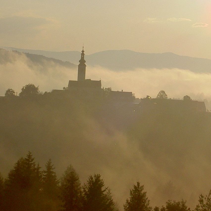 Eine neblige Morgenlandschaft mit einem hohen Turm und Bergen in der Ferne, getaucht im goldenen Licht der aufgehenden Sonne.