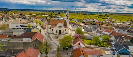 Ein Luftbild einer Stadt mit einer Kirche, Häusern und Windrädern in der Ferne. Bäume und Pflanzen umgeben das Gebiet, und eine Straße mit einem gehender Person.
