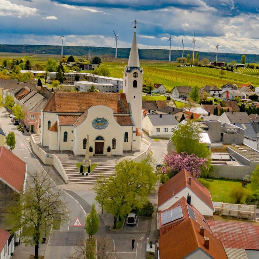 Ein Luftbild einer Stadt mit einer Kirche, Häusern und Windrädern in der Ferne. Bäume und Pflanzen umgeben das Gebiet, und eine Straße mit einem gehender Person.