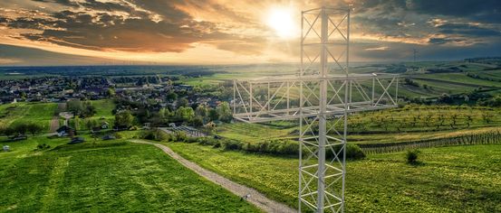 Ein weißer Metallkreuz steht auf einer grünen Anhöhe, umgeben von einer ländlichen Landschaft mit Häusern und einem Sonnenuntergang im Hintergrund.