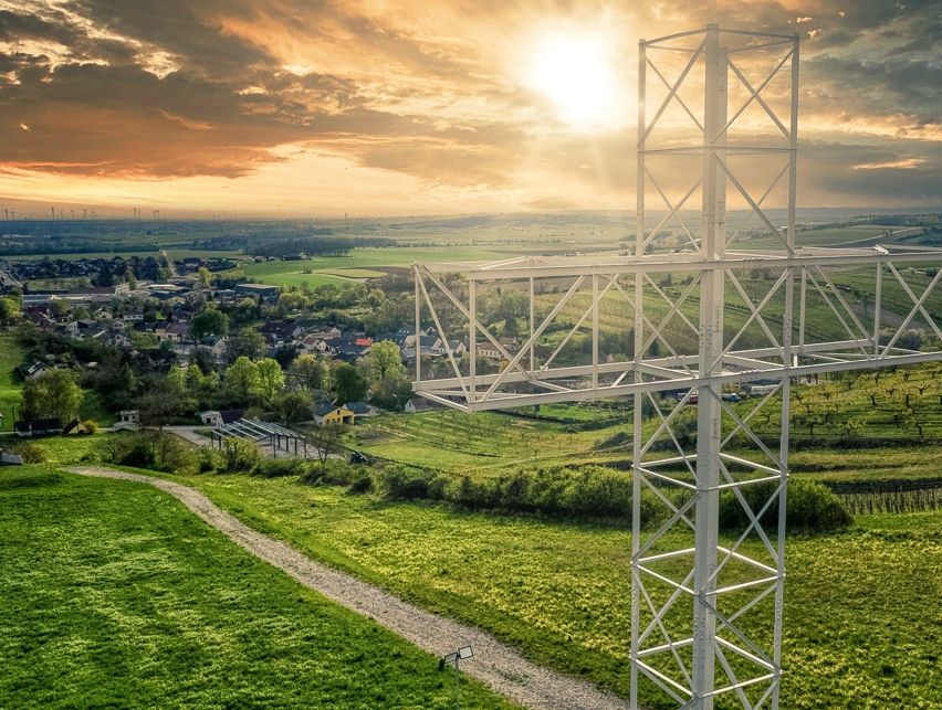 Ein weißer Metallkreuz steht auf einer grünen Anhöhe, umgeben von einer ländlichen Landschaft mit Häusern und einem Sonnenuntergang im Hintergrund.