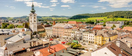 Luftaufnahme einer Stadt mit vielen Gebäuden, einem Uhrturm, grünen Feldern und Bergen in der Ferne.