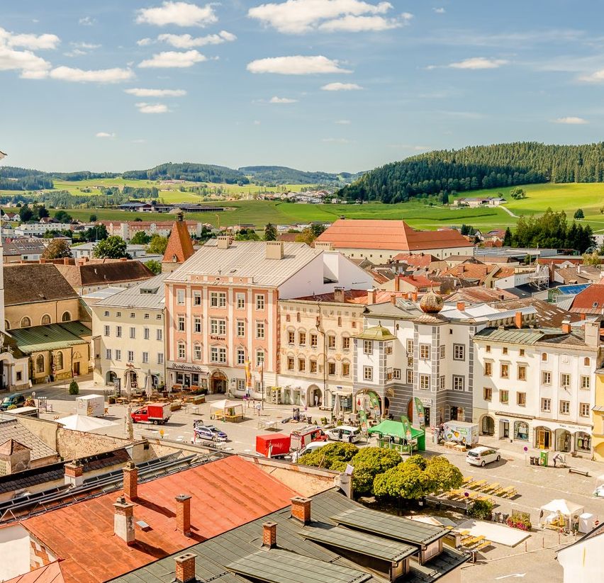Luftaufnahme einer Stadt mit vielen Gebäuden, einem Uhrturm, grünen Feldern und Bergen in der Ferne.