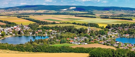 Luftaufnahme eines Dorfes umgeben von üppigen grünen Feldern und Wäldern, mit einem großen weißen Kreidehügel in der Ferne.
