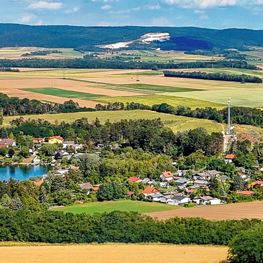 Luftaufnahme eines Dorfes umgeben von üppigen grünen Feldern und Wäldern, mit einem großen weißen Kreidehügel in der Ferne.