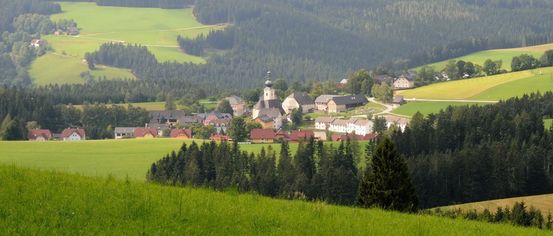 Ein ländliches Dorf, umgeben von üppigen grünen Feldern und dichten Wäldern. Die Häuser sind in verschiedenen Farben gestrichen, mit einem Kirchturm in der Mitte.