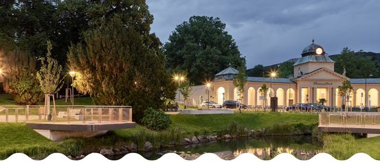 Eine Abendszene mit einem Gebäude, möglicherweise einem Gartenpavillon, beleuchtet von Straßenlaternen. Bäume und Pflanzen umgeben das Gebiet, und ein Gewässer spiegelt die Lichter wider. Autos sind davor geparkt.