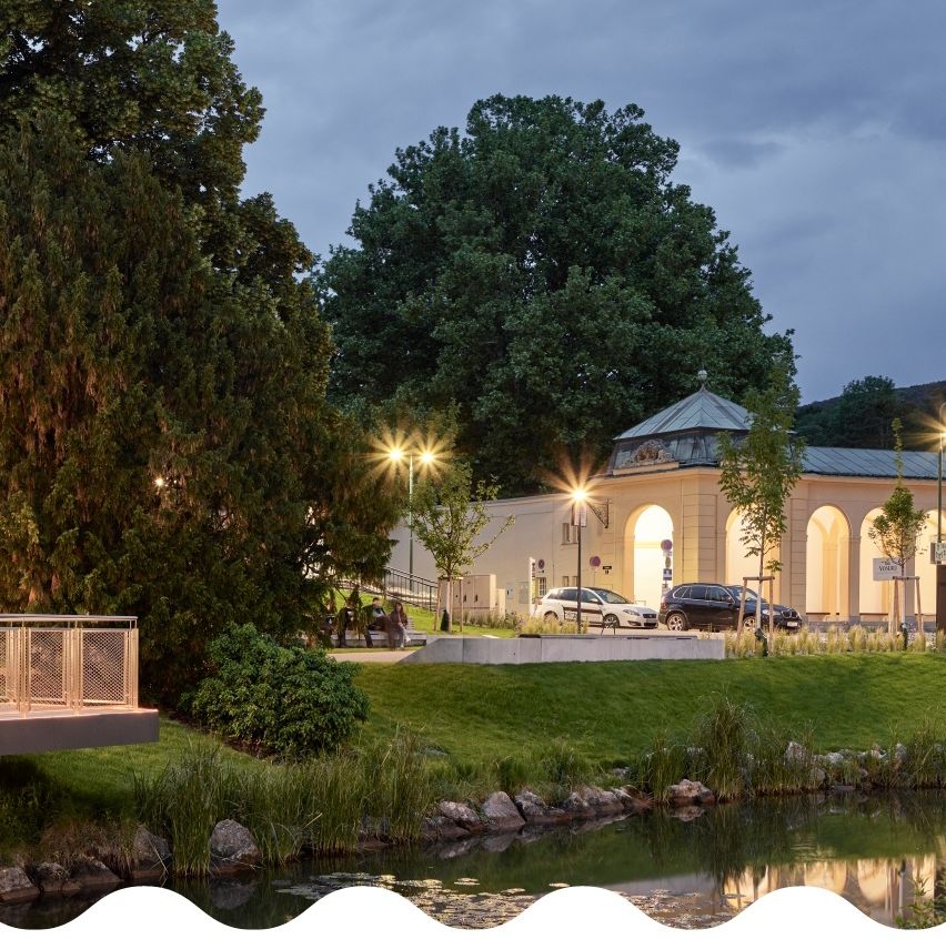 Eine Abendszene mit einem Gebäude, möglicherweise einem Gartenpavillon, beleuchtet von Straßenlaternen. Bäume und Pflanzen umgeben das Gebiet, und ein Gewässer spiegelt die Lichter wider. Autos sind davor geparkt.