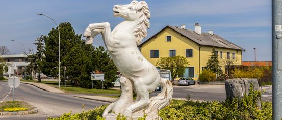 Eine weiße Pferdestatue steht in einem Busch mit einem gelben Haus und einer Straße dahinter. Der Himmel ist klar und es gibt eine Straßenlaterne links.