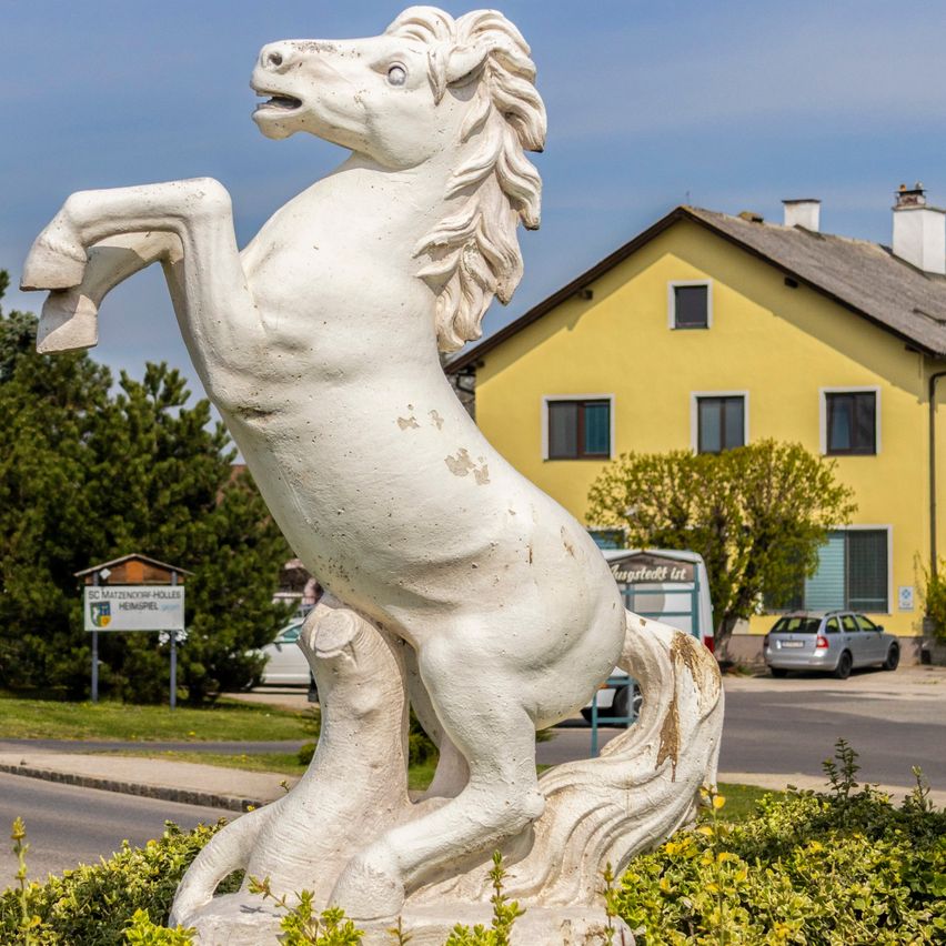 Eine weiße Pferdestatue steht in einem Busch mit einem gelben Haus und einer Straße dahinter. Der Himmel ist klar und es gibt eine Straßenlaterne links.