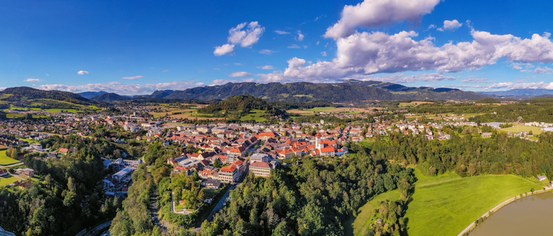 Luftaufnahme einer Stadt umgeben von üppigem Grün und Bergen unter einem teilweise bewölkten blauen Himmel.