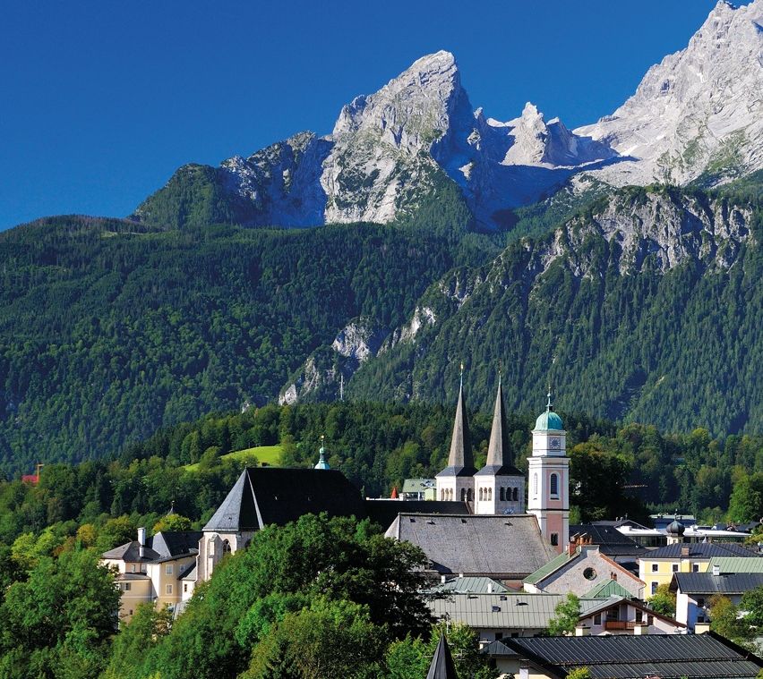 Eine von Bergen umgebene Stadt, mit einem sichtbaren Kirchturm und einem klaren blauen Himmel darüber. Bäume und Häuser säumen die Landschaft, und ein Holzzaun begrenzt die Szene.