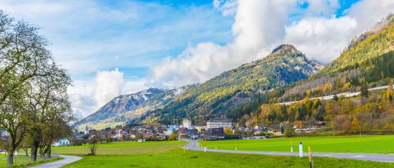 Ein Dorf in einem Tal, umgeben von üppigen grünen Feldern, Bergen und einem klaren blauen Himmel mit verstreuten Wolken.
