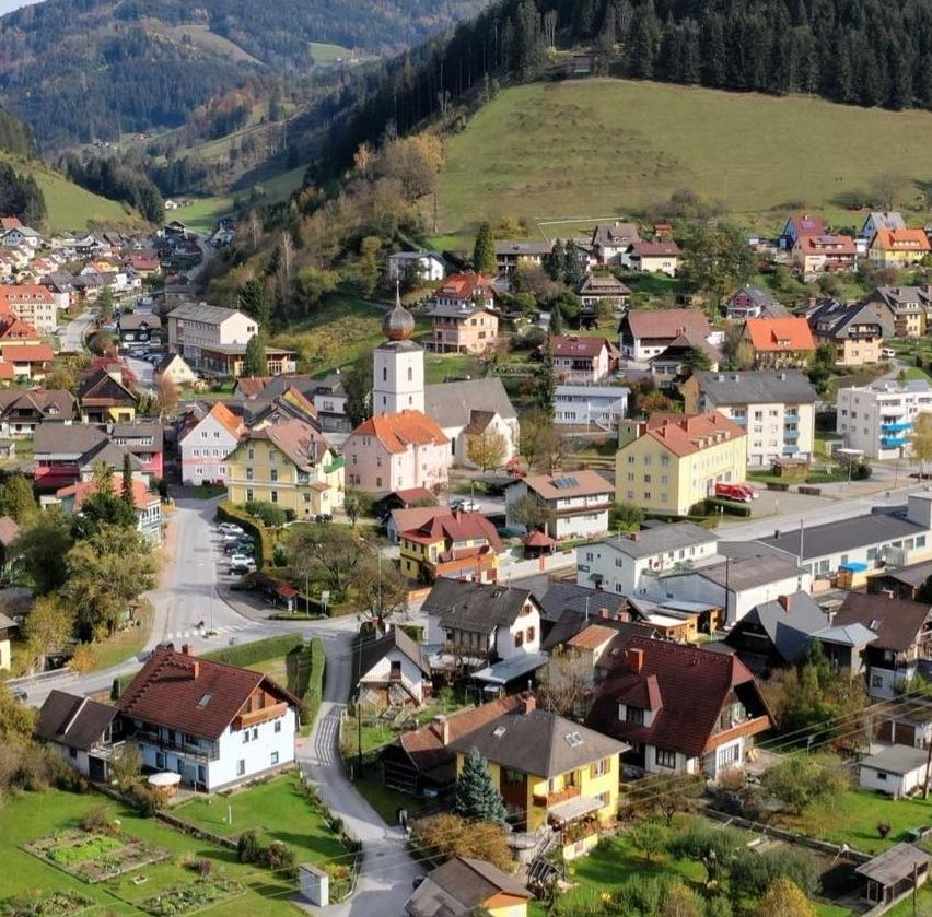 Luftaufnahme eines kleinen Dorfes in den Bergen, mit Häusern in verschiedenen Farben und einer Kirche in der Mitte.