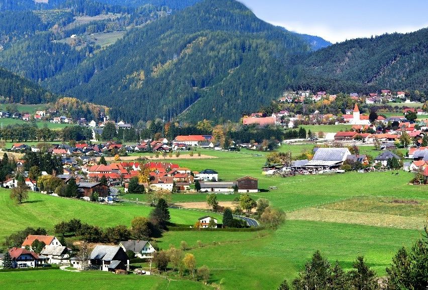 Ein Luftbild eines kleinen Dorfes, umgeben von üppigen grünen Feldern und Bergen. Gebäude mit roten Dächern säumen die Landschaft.